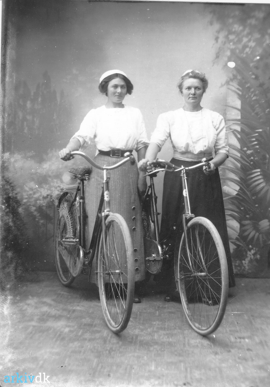 arkiv.dk | To damer, iklædt hvide bluser, og lange nederdele. Står med cykler.
